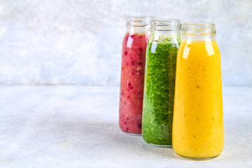 Green, yellow, purple smoothies in bottles of berries, greens, oranges on a white board on a gray table.