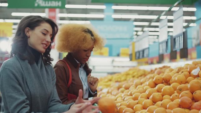 Two cute young women chooses orange in supermarket walk smile food shopping fruit black working consumer family buying fresh grocery market portrait mixed customers slow motion close up