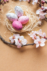 Obraz na płótnie Canvas View of a straw nest with colorful pink and white freckled Easter eggs and tree branches blooming with white flowers on brown cardboard background, Happy Easter background