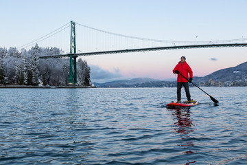 Adventurous man on a Standup Paddle Board is padding near Lions Gate Bridge during a vibrant winter sunrise. Taken in Vancouver, British Columbia, Canada.