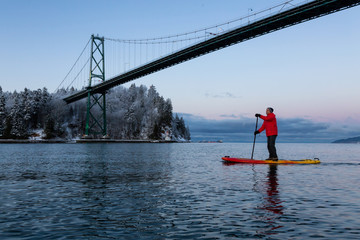 Naklejka premium Adventurous man on a Standup Paddle Board is padding near Lions Gate Bridge during a vibrant winter sunrise. Taken in Vancouver, British Columbia, Canada.