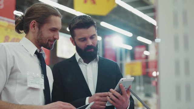 Smiling seller consultant talk with young beard man use hold touch phone in tech store business hand shopping electronics technology internet mall slow motion portrait close up handsome attractive