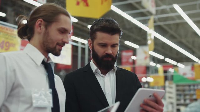 Handsome seller consultant talk with young beard man use hold touch tablet computer in tech store business hand shopping electronics technology internet mall slow motion portrait close up attractive