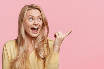 Indoor shot of amazed blonde woman has overjoyed expression, indicates with excited look aside,...