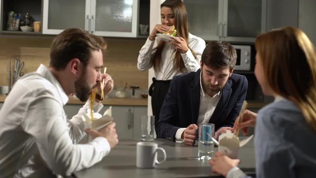 Portrait of positive people women and men having lunch break at work, eating noodles from boxes using chopsticks and drinking tea sitting at kitchen table, slow motion