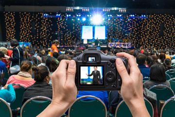 Hand holding the camera taking photograph to the businessman on the stage over the Rear view of...