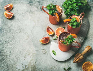 Blood orange Moscow mule alcohol cocktails with fresh mint leaves and ice in copper mugs on board over grey concrete background, copy space
