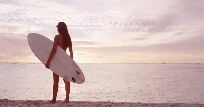 Surfer woman on beach vacation travel going surfing walking with surfboard at sunset on tropical beach. Surfing girl at ocean sunset. Female bikini woman by water livinmg healthy active lifestyle.