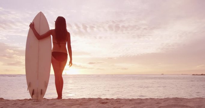 Travel vacation surfing - surfer woman looking at ocean beach sunset. Sexy woman in bikini going surfing looking at water standing with surfboard living healthy active lifestyle. RED EPIC SLOW MOTION.