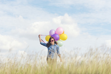 Happy cheerful woman holding colorful of balloons wear sunglasses on green meadow with cloudy blue sky. Travel Concept