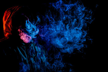 A young man in a black hood smokes and breathes out the colored blue and red smoke from the vape on...