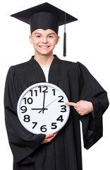 Portrait of a graduate teen boy student in a black graduation gown with hat, holding big clock - isolated on white background. Child back to school, educational and time concept.