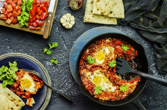 Middle Eastern traditional food; Shakshuka skillet served with pita bread and placed with fresh chopped vegetables. Top view with close-up