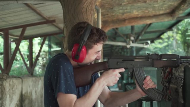 Slow motion: white millennial man smiling while firing ak 47 automatic assault rifle at gun range in Cu Chi Tunnels, Vietnam