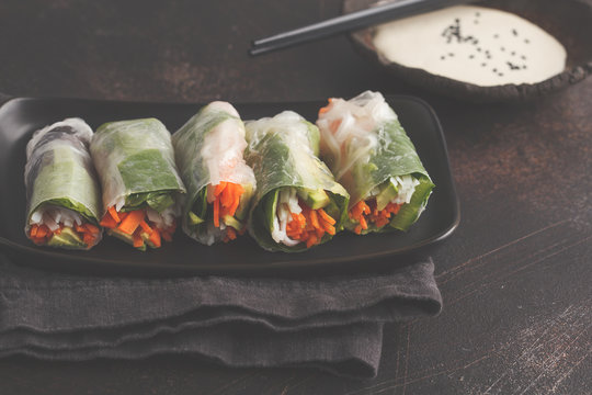 Fresh handmade vegan asian spring rolls with rice noodles, avocado, carrots and tahini dressing on black dish, dark background.
