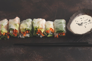Fresh handmade vegan asian spring rolls with rice noodles, avocado, carrots and tahini dressing on dark background, copy space.