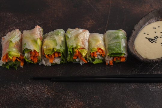 Flat-lay of vegan spring, summer rice paper rolls with vegetables: rice noodles, avocado, carrots and tahini dressing on dark background, copy space.