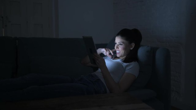 lateral pan shot of young beautiful happy and relaxed latin woman lying on home couch late night using digital device laptop tablet pad watching internet movie 