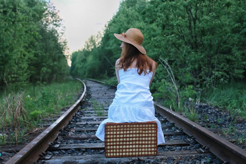 girl in a white sundress and wicker suitcase walking on rails