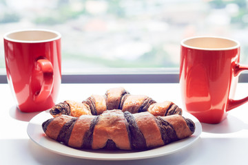 Red cup of morning coffee and croissant on a white wooden table.