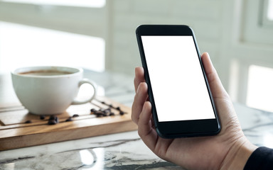 Mockup image of hands holding black mobile phone with blank white screen with a cup of coffee in cafe