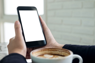 Mockup image of hands holding black mobile phone with blank white screen with a cup of coffee in cafe