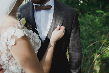 Beautiful wedding couple in the forest. The bride touches the groom in bow tie tenderly. Wedding buttonhole and checkered suit in Great Gatsby style. Rustic and stylish.