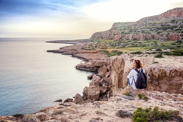 Foto op Canvas A stylish young woman traveler watches a beautiful sunset on the rocks on the beach, Cyprus, Cape Greco, a popular destination for summer travel in Europe © olezzo