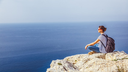 A girl traveler sits on a rock and admires the blue boundless sea, freedom, travel, unity with nature,