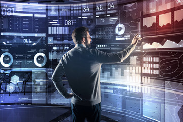 Responsible programmer. Calm clever experienced programmer standing with one hand on the hip and carefully touching the icon on a futuristic transparent device