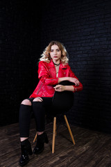 Studio portrait of blonde girl in red leather jacket posed on chair against brick wall.