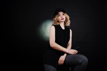 Studio portrait of blonde girl in black wear and cap with mobile phone at hand against dark background.