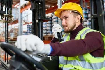 Waist up portrait of young man sitting in forklift and using walkie-talkie while moving goods in...