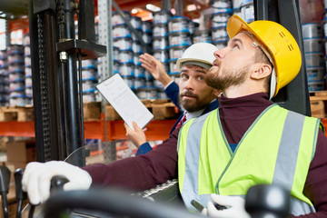 Side view portrait of young man sitting in forklift car looking up and moving goods with supervisor...