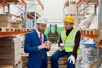 Portrait of two warehouse workers taking break sitting on pellets drinking coffee and chatting