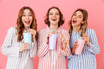 Fototapeta premium Three beautiful young girls 20s wearing colorful striped pyjamas smiling and drinking cold soda from paper cups during happy sleepover, isolated over pink background