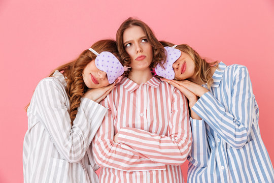 Three friendly young girls 20s wearing colorful striped pyjamas and sleeping masks sleeping while playing around during girlish sleepover, isolated over pink background