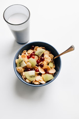 Delicious crispy cornflakes with kiwi pieces and cranberries in bowl, glass of milk on white background, closeup, healthy breakfast