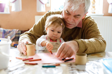 Cute little baby toddler girl and handsome senior grandfather painting with colorful pencils at home. Grandchild and man having fun together - obrazy, fototapety, plakaty