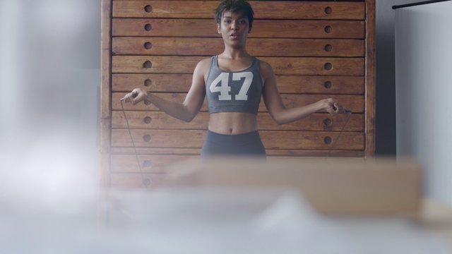 african american woman makes a jump rope routine in sunny loft for healthy life. front view throught reflecting glasses