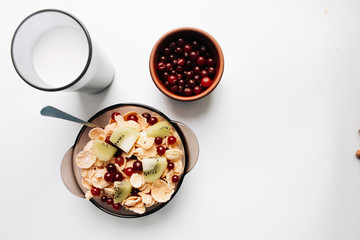 Delicious crispy cornflakes with kiwi pieces and cranberries in bowl, glass of milk on white background, closeup, healthy breakfast