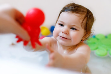 Cute adorable baby girl taking foamy bath in bathtub. Toddler playing with bath rubber toys.