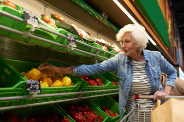 Portrait of modern senior woman with shopping cart  choosing fruits and vegetables in supermarket while enjoying grocery shopping, copy space