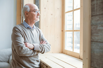 Pensive mood. Thoughtful attractive senior man gazing through window while crossing hands and staying