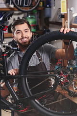 handsome young man in apron fixing bicycle and smiling at camera in workshop