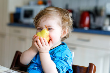 Cute adorable toddler girl eating fresh pear . Hungry happy baby child of one year holding fruit.