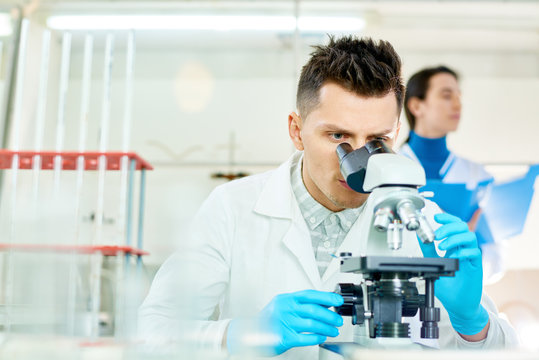 Handsome young scientist with stylish haircut using microscope while working on ambitious project, interior of modern laboratory on background