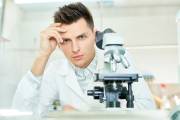 Naklejka na ściany i meble Portrait shot of handsome young researcher with stylish haircut sitting at laboratory bench and looking at camera, modern microscope on foreground