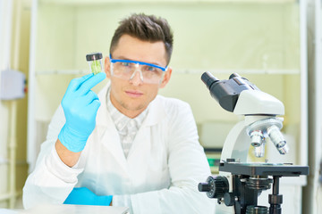 Waist-up portrait of confident young researcher wearing safety goggles and white coat sitting at desk of modern lab with GMO lettuce sample in hand and looking at camera.