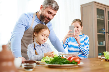 Portrait of smiling father teaching little girl cooking in kitchen while another daughter taking photo of family scene